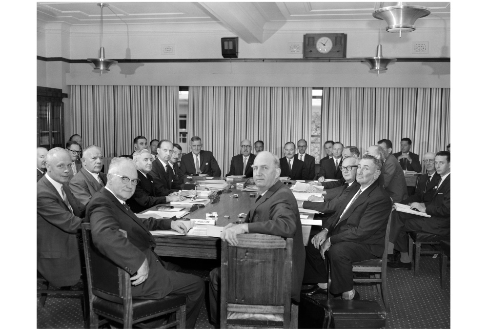 Black and white photograph of a large group of older men in suits seated around a table in a conference room. They are all looking at the camera. In the image are (from the far head of the table, clockwise) federal Ministers Hugh Roberton, Paul Hasluck; H Winston Noble from Queensland, Charles Collier Perkins and SG Middleton from WA; GC Smith from Tasmania; and Glen Pearson from South Australia, among others. NAA: A1200, L37460.