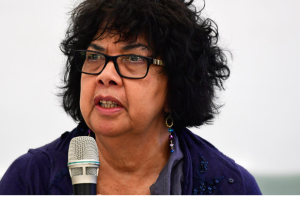 Portrait photograph of Dr Dawn Casey, speaking with a microphone in front of her. She is wearing glasses and a purple woollen top.