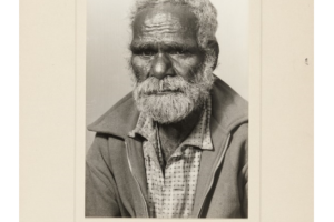 Photo of Norman Bilson, also known as Walaru. Bilson has white hair, a short beard and wrinkles. He has a serious expression. He is dressed in a button-up shirt and zip-up jacket. The photo is mounted on card and there is a handwritten caption beneath it. The caption reads: ‘Norman Bilson (Walaru) Aborigine Station Hand, suffers from cataracts, but is still in request for his skilled work. W.A. 1961. Age 66 in 1960.’ 