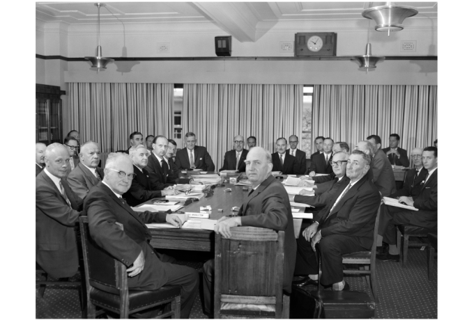 Black and white photograph of a large group of older men in suits seated around a table in a conference room. They are all looking at the camera. In the image are (from the far head of the table, clockwise) federal Ministers Hugh Roberton, Paul Hasluck; H Winston Noble from Queensland, Charles Collier Perkins and SG Middleton from WA; GC Smith from Tasmania; and Glen Pearson from South Australia, among others. NAA: A1200, L37460.